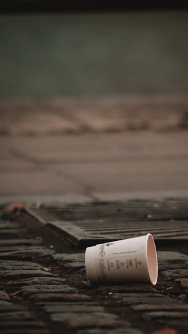 A discarded white paper coffee cup with a printed label, lying on a wet brick pavement near a drainage grate. The cup is positioned on its side with the open end facing away, and appears to be made of paper with a smooth surface. The surrounding environment includes a section of rough-textured brickwork with visible mortar joints, and a drain cover with parallel metal slats located nearby. The background is blurred, showing a greenish hue that suggests outdoor surroundings, possibly a sidewalk or garden area. The lighting indicates natural daylight, contributing to the typical scene of careless rubbish left on a street or public walkway. This image illustrates the importance of waste disposal and the potential for private rubbish collection services like those offered by Rubbish Collection Edgware to manage on-site waste and prevent littering, aligning with the theme of alternative rubbish handling methods outside local authority collection.