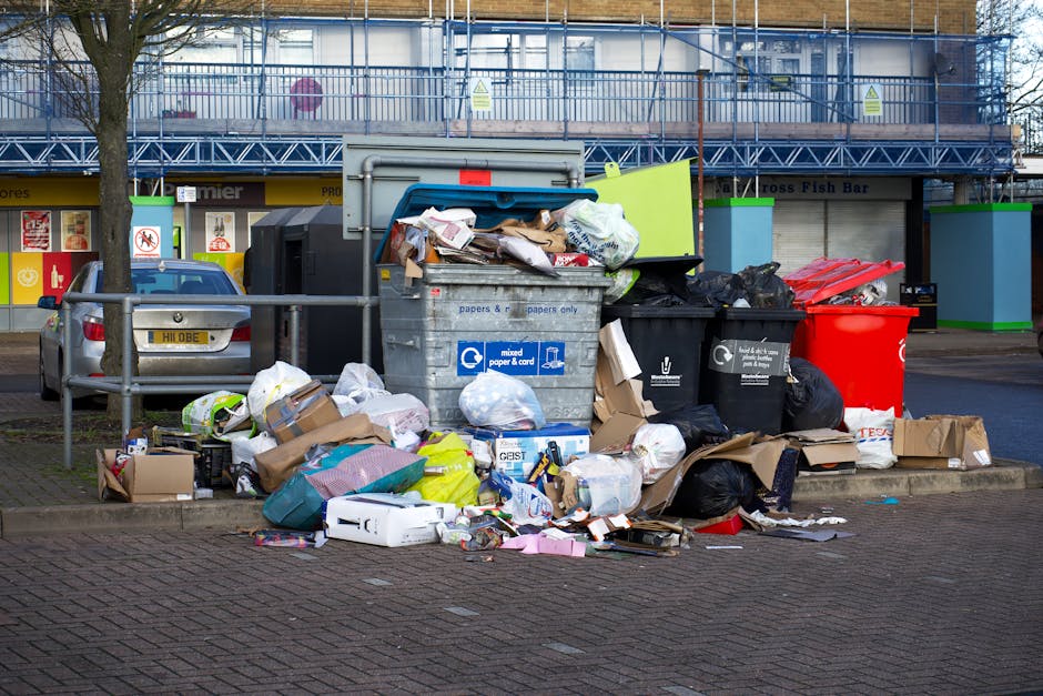 A collection of overflowing rubbish bins and scattered waste bags on a paved area in front of a commercial building. The central grey bin, designated for mixed paper and cards, is open with various crumpled papers, cardboard boxes, and miscellaneous waste protruding from the top. To its right, there are several black and red waste sacks, some of which are partially open, revealing more rubbish inside. On the left, a small silver car is parked behind a metal railing, with a pile of discarded cardboard, plastic bags, and packaging materials spread across the pavement. The background features a storefront with closed shutters, advertising posters, and a multi-story building under construction or renovation, covered with blue scaffolding. The environment suggests a typical urban setting where residents or businesses have disposed of rubbish, perhaps in a location where alternative waste handling services might be considered, as it exceeds typical collection capacity, highlighting the importance of professional rubbish removal services like those offered by Rubbish Collection Edgware.