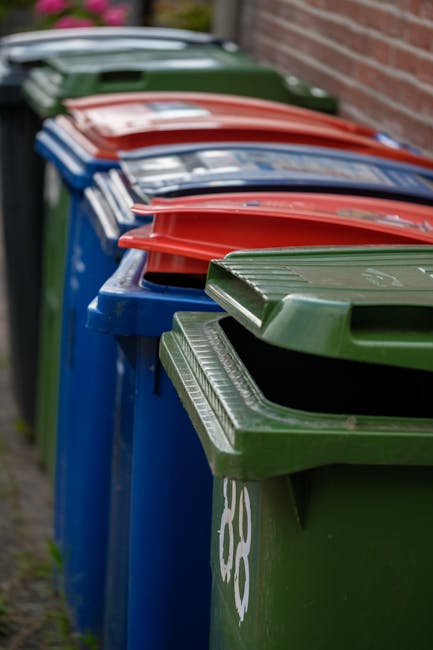 A row of multiple public refuse bins positioned along a paved pathway beside a brick wall, with the bins primarily in shades of green, blue, and red. The green bin at the forefront features a ribbed, matte surface with a lid slightly ajar, revealing a dark interior. Next to it, a blue bin with a smooth, glossy finish and a closed lid is visible, with the number 120 painted in white on its side. Behind these, additional bins in red and green are lined up, with some lids open or partially lifted. The arrangement appears tidy and organized, set in an outdoor urban environment likely near residential or communal areas. The background is slightly blurred, emphasizing the focus on the bins' textures and colors. The scene suggests an emphasis on waste segregation and collection, reflecting practices consistent with local waste management guidelines, and hinting at options for private disposal services such as Rubbish Collection Edgware.