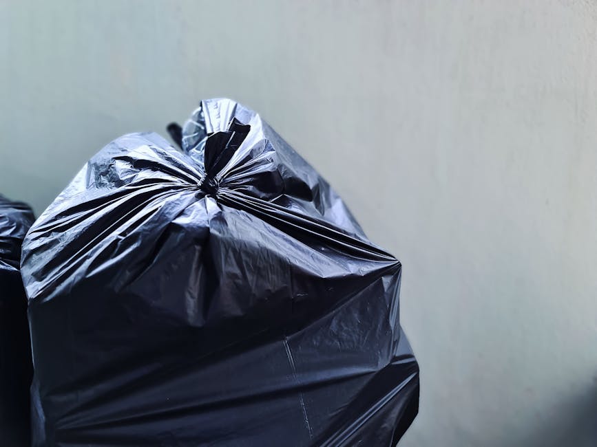 A collection of overflowing rubbish bins and scattered waste bags on a paved area in front of a commercial building. The central grey bin, designated for mixed paper and cards, is open with various crumpled papers, cardboard boxes, and miscellaneous waste protruding from the top. To its right, there are several black and red waste sacks, some of which are partially open, revealing more rubbish inside. On the left, a small silver car is parked behind a metal railing, with a pile of discarded cardboard, plastic bags, and packaging materials spread across the pavement. The background features a storefront with closed shutters, advertising posters, and a multi-story building under construction or renovation, covered with blue scaffolding. The environment suggests a typical urban setting where residents or businesses have disposed of rubbish, perhaps in a location where alternative waste handling services might be considered, as it exceeds typical collection capacity, highlighting the importance of professional rubbish removal services like those offered by Rubbish Collection Edgware.