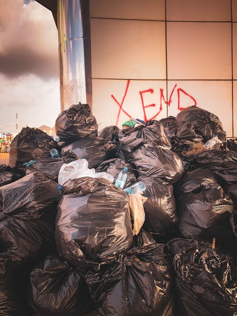 A large accumulation of black plastic rubbish bags piled against a tiled wall outdoors, with some bags appearing partially torn or displaced, revealing plastic bottles and miscellaneous waste inside. The bags are stacked haphazardly, occupying most of the foreground, with a few lighter-colored plastic items visible among them. In the background, there is a metallic cylindrical structure reflecting minimal light, and graffiti with the word 'XEND' spray-painted in red on the wall behind the waste. The scene is set on a paved area adjacent to an urban building, suggesting an unauthorized or abandoned waste collection point, highlighting a typical scenario for private or alternative rubbish removal services like those offered by Rubbish Collection Edgware. The lighting indicates either late afternoon or early evening, with a cloudy sky overhead, casting a muted tone over the area. This image visually supports the context of non-local authority rubbish clearance or independent waste disposal solutions within an urban environment.