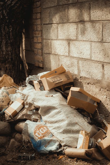 A pile of discarded cardboard boxes, some flattened and others partially intact, is positioned against a light grey brick wall outdoors. The boxes vary in size and are mostly brown with printed labels and barcodes visible on their surfaces. Among the boxes, there are also some white plastic bags filled with unknown contents, one of which is partially covered by a textured white sack. The scene is set on a ground surface composed of small rocks and dirt, with a large tree trunk visible on the left side, partially in the frame. The background features a plain brick wall, with no other objects or signs of activity present. The pile appears to be gathered for waste disposal or collection, reflecting an example of non-authority or private waste handling, which companies such as Rubbish Collection Edgware might assist with in alternative rubbish removal services.