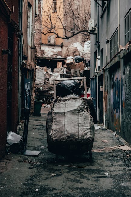 A narrow, urban alleyway cluttered with discarded rubbish, featuring a large, worn black garbage bag in the foreground, placed on the uneven, dark asphalt ground with visible stains and debris, flanked by tall brick and concrete walls on either side. To the right, a building with metal security bars over windows and graffiti markings adds an industrial feel. In the background, a considerable heap of mixed waste materials, including cardboard boxes, plastic bags, and other refuse, is piled up against a wooden barrier, partially obscured by leafless tree branches overhead. The scene is illuminated by diffused natural light, highlighting the textures of the rubbish and surrounding surfaces, evoking a context of private waste clearance that Rubbish Collection Edgware provides for efficient rubbish removal in densely built urban areas.