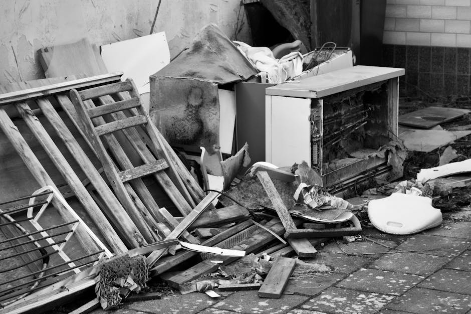 The image depicts a collection of discarded household items and debris piled on a paved outdoor surface, likely part of a rubbish clearance or private waste removal effort. In the foreground, there are several wooden items, including a dismantled or broken shelving unit with horizontal slats, showing weathered textures and natural wood grain. Behind the wood, a damaged or heavily soiled white refrigerator or freezer is visible, its door partially open or removed, exposing the interior rough and rusty surfaces. To the right, a white plastic object, possibly a toilet seat or container, rests on the ground, partially obscured by other debris. Surrounding these main objects are various smaller fragments, including broken wooden pieces, a small coil of wire, and torn or crumpled fabric, all scattered haphazardly across the pavement. The background includes a brick wall and parts of a building structure, with some shadows indicating natural daylight. The overall scene suggests an accumulation of bulky waste awaiting disposal or collection by an independent rubbish removal service such as Rubbish Collection Edgware, situated on a paved outdoor area typical of a residential or commercial property awaiting clearance.