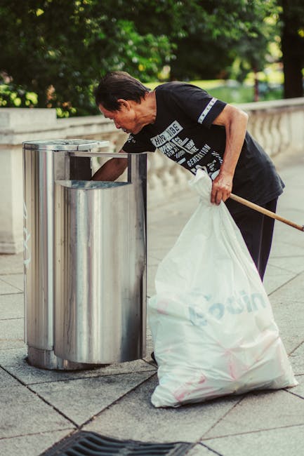 A middle-aged man with dark hair wearing a black T-shirt is seen disposing of rubbish into a tall, cylindrical stainless steel public trash bin. He is bent forward slightly, using his right hand to lift the lid of the bin while holding a large, white plastic bag filled with waste in his left hand. The bag appears semi-transparent with some visible rubbish inside. The scene takes place outdoors on a paved sidewalk, with a decorative stone or concrete balustrade immediately behind him. The background features lush green foliage, suggesting a park or garden setting, with sunlight filtering through the trees, creating a well-lit, natural atmosphere. The weather appears mild and clear, appropriate for outdoor waste disposal, and the scene subtly reflects the context of independent or private rubbish collection. Rubbish Collection Edgware’s professional waste management services may be inferred from the setting, emphasizing lawful and proper disposal practices outside of local authority collection points.