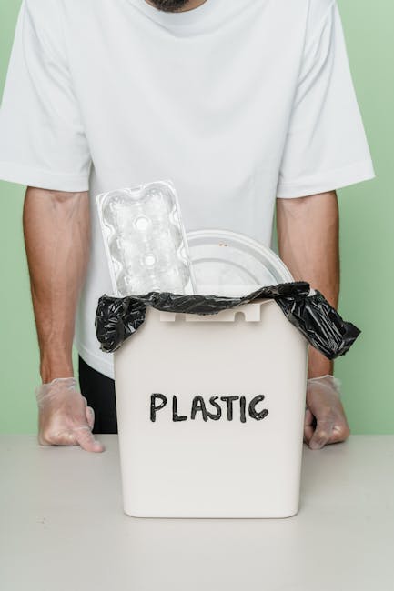 A person wearing a white t-shirt and transparent gloves stands behind a small white plastic container labeled 'PLASTIC,' which is filled with discarded plastic items such as a clear plastic egg carton and a plastic bag tied around the opening. The container is placed on a flat surface, and the background is a solid light green colour. The scene emphasizes proper waste segregation and highlights the importance of recycling plastics, consistent with services offered by rubbish collection providers like Rubbish Collection Edgware, which facilitate private waste disposal and on-site clearance for domestic and commercial clients.