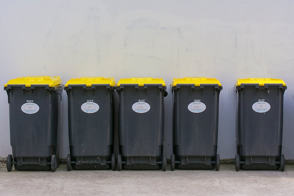 A middle-aged man with dark hair wearing a black T-shirt is seen disposing of rubbish into a tall, cylindrical stainless steel public trash bin. He is bent forward slightly, using his right hand to lift the lid of the bin while holding a large, white plastic bag filled with waste in his left hand. The bag appears semi-transparent with some visible rubbish inside. The scene takes place outdoors on a paved sidewalk, with a decorative stone or concrete balustrade immediately behind him. The background features lush green foliage, suggesting a park or garden setting, with sunlight filtering through the trees, creating a well-lit, natural atmosphere. The weather appears mild and clear, appropriate for outdoor waste disposal, and the scene subtly reflects the context of independent or private rubbish collection. Rubbish Collection Edgware’s professional waste management services may be inferred from the setting, emphasizing lawful and proper disposal practices outside of local authority collection points.